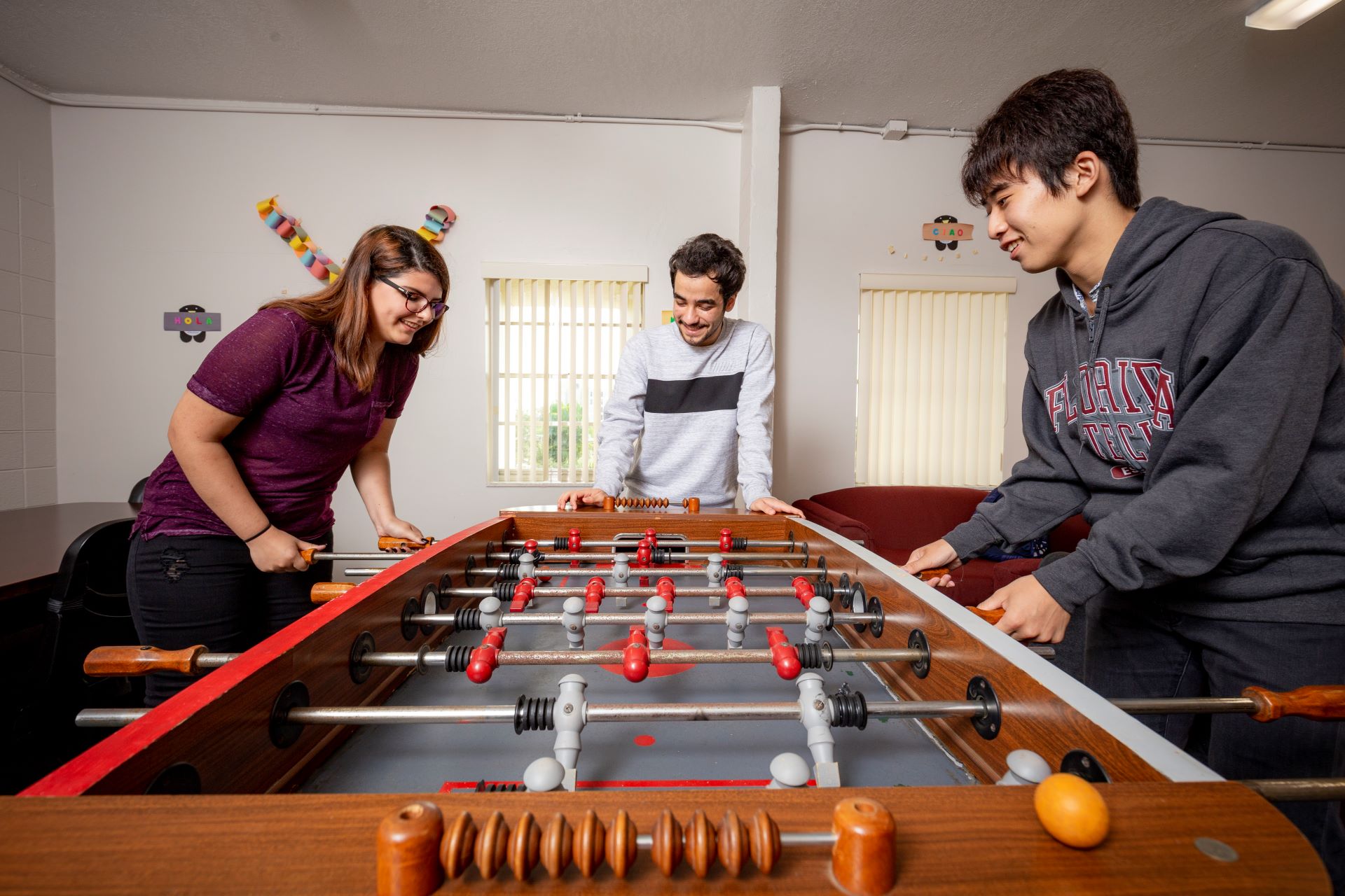 Three students, two men and one woman, playing foosball in a brightly lit room with decorations on the walls